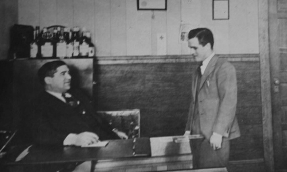 Old photo of two men, one standing, and talking